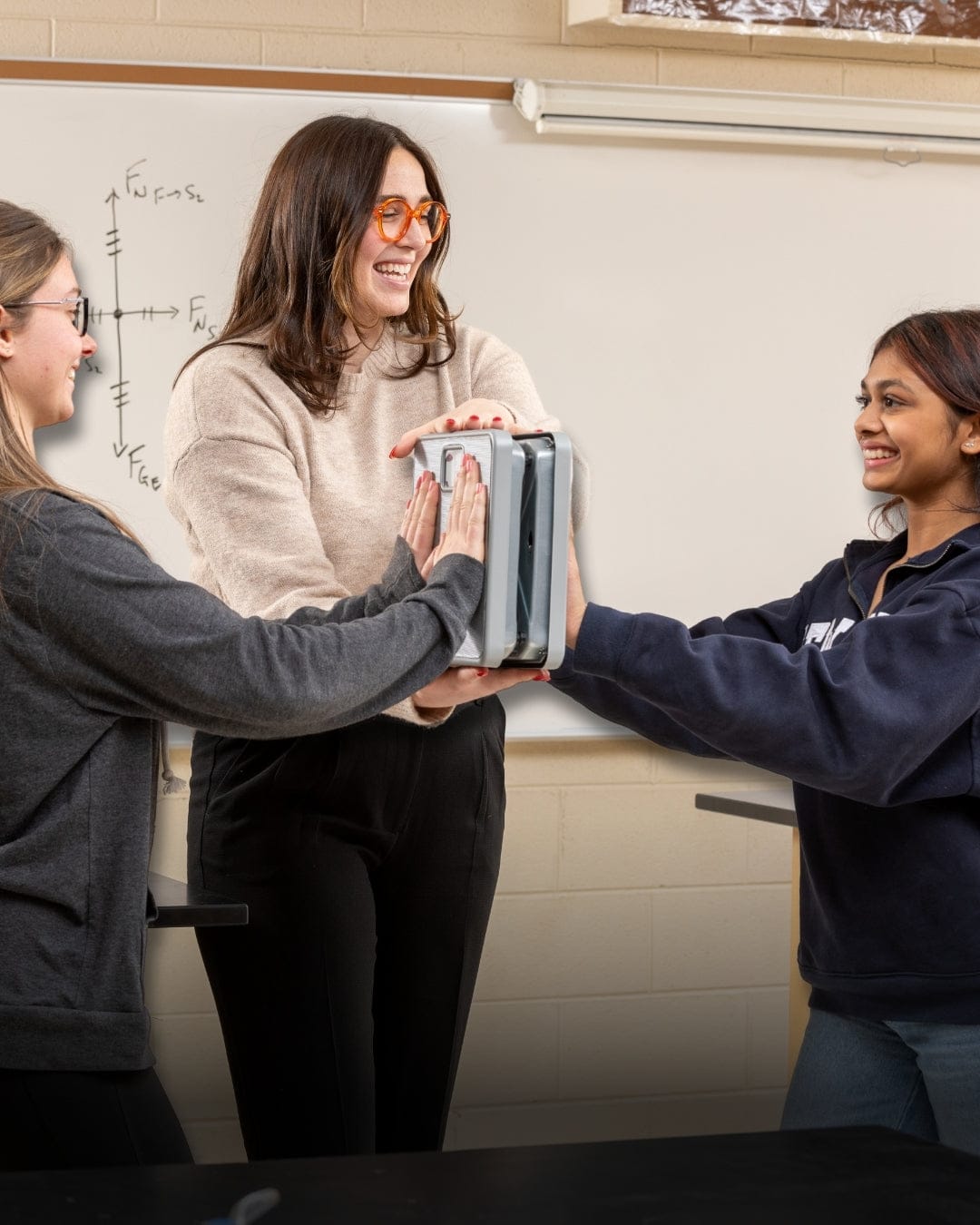 A teacher with two students in a classroom setting, two students pushing against bathroom scales to show force pairs