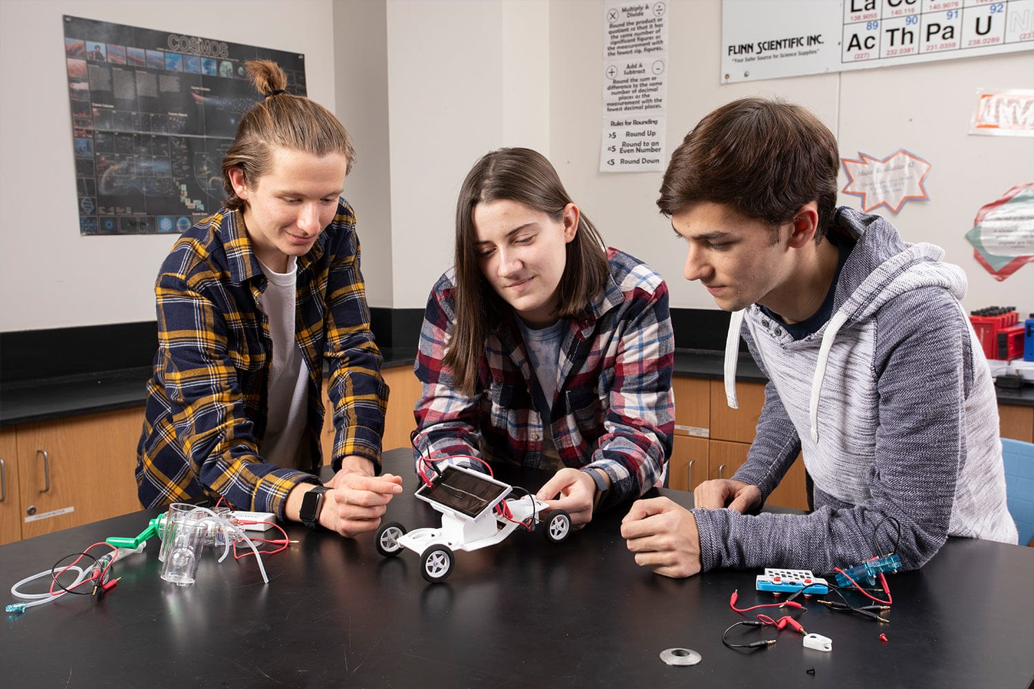 Three students working on a science project with a small hydrogen fuel cell powered vehicle on a table in a classroom.